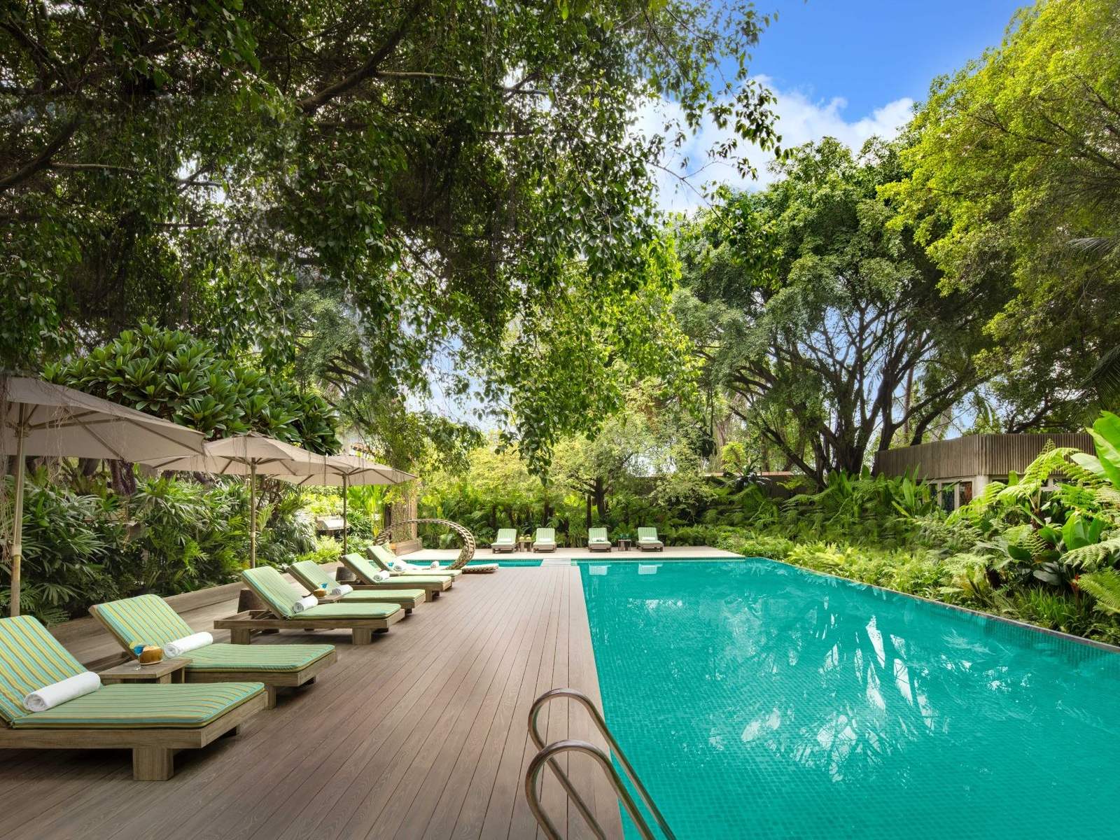 Long turquoise pool lined with green loungers and parasols beside a lawn and tall trees