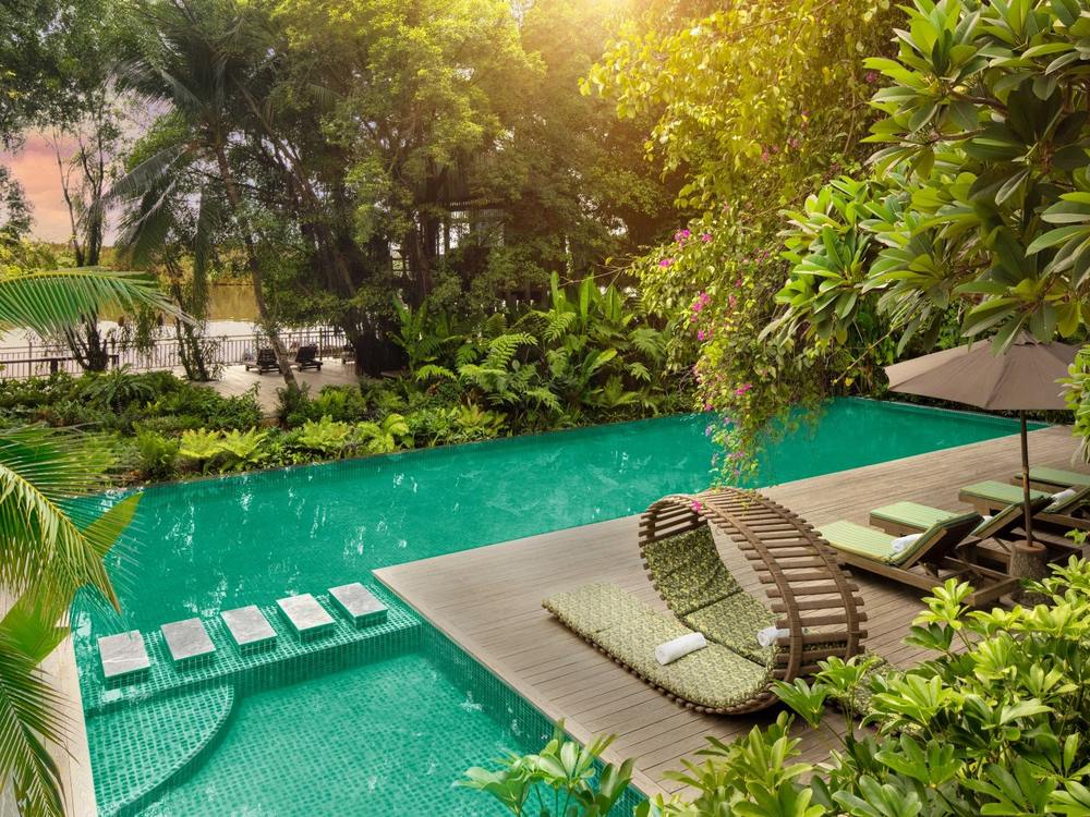 Long turquoise pool with stepping stones and a round woven daybed, framed by tropical planting with the river beyond