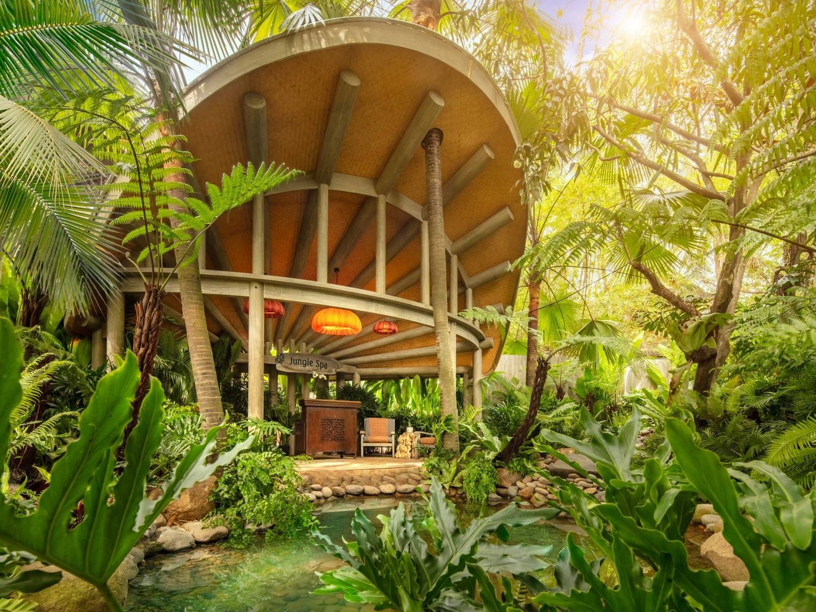 Round timber pavilion with a curved roof half hidden in sunlit tropical trees