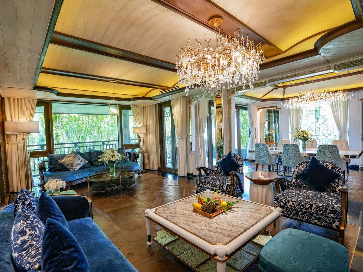 Lounge with blue velvet sofas and a crystal chandelier, windows open onto treetops