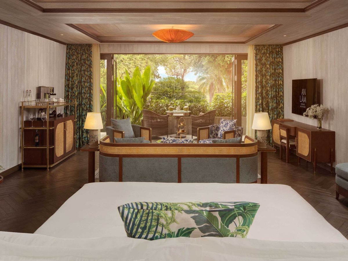 Large bed facing wide windows onto garden and terrace, with rattan furniture and a drinks trolley