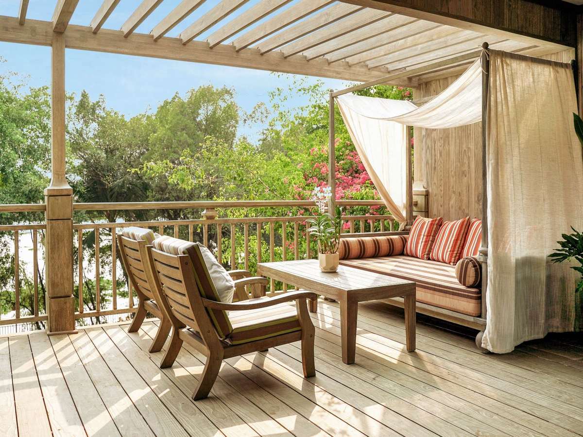 Covered timber terrace with wooden armchairs and a draped daybed looking out over trees