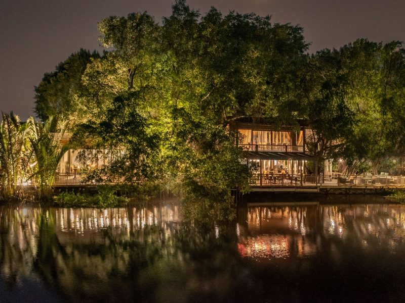 Riverside building lit up at night among large trees, reflected in the dark river