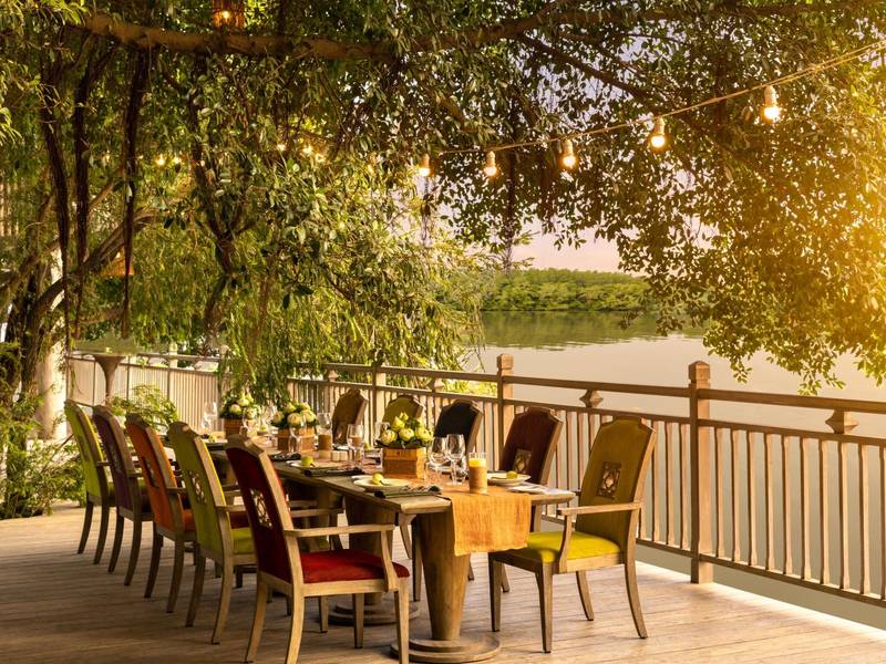 Long dinner table with colourful chairs on a wooden deck under banyan trees beside the river
